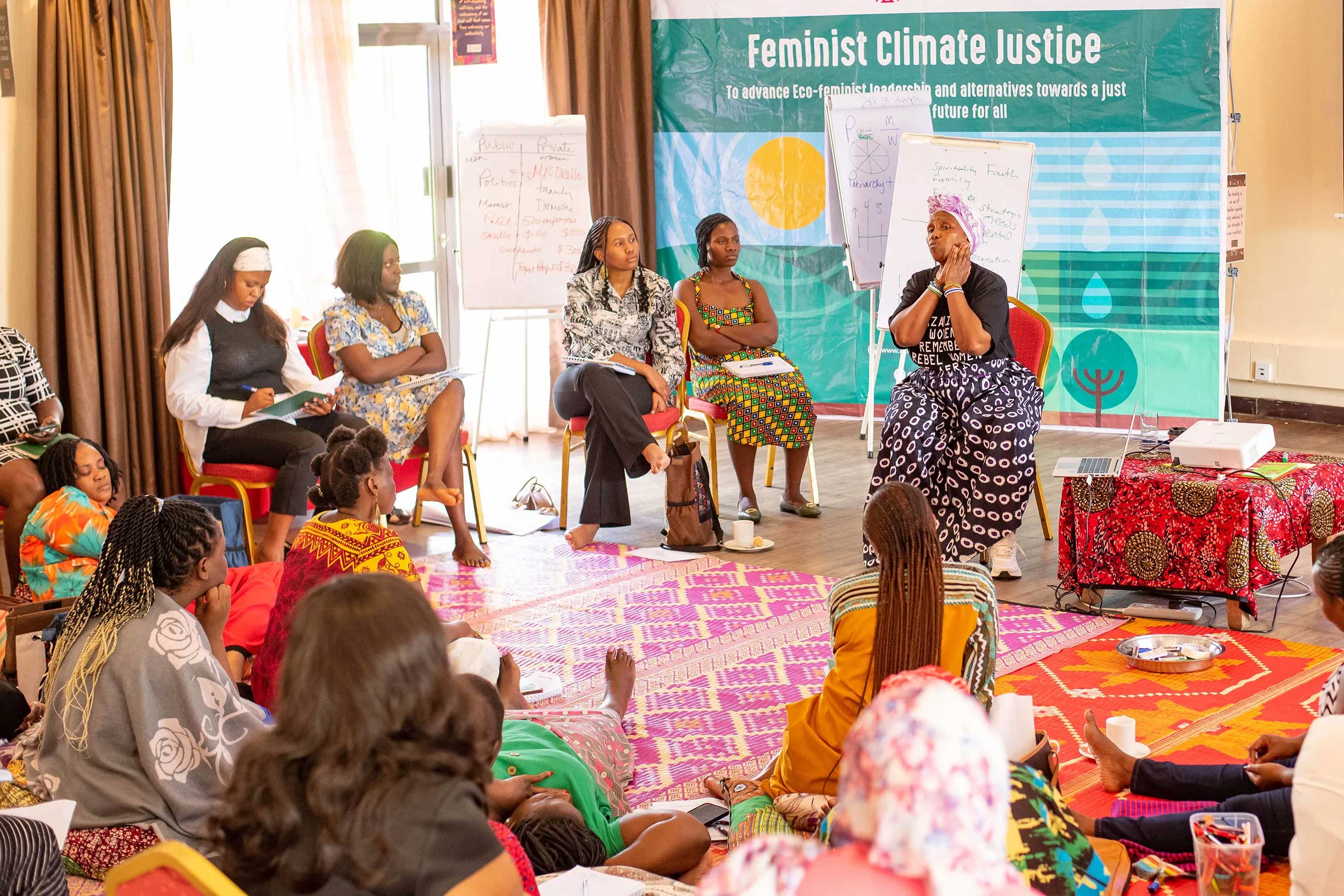 A group of people, mostly women, are gathered for a conference or workshop. Several are seated in chairs on a stage while the audience sits on the floor. A banner behind the stage reads "Feminist Climate Justice."