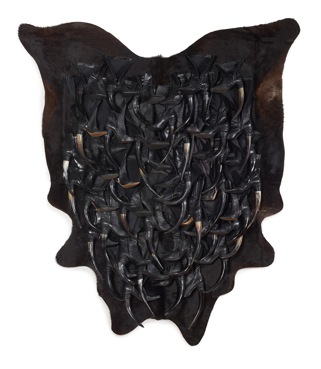 A wall-mounted hanging sculpture of leather boots with animal horn heels adhered to an animal hide.