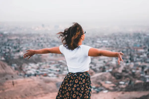 A person with a white shirt and dark skirt with their back to the camera stands on a hill with out-stretched arms overlooking a sprawling, hazy city.