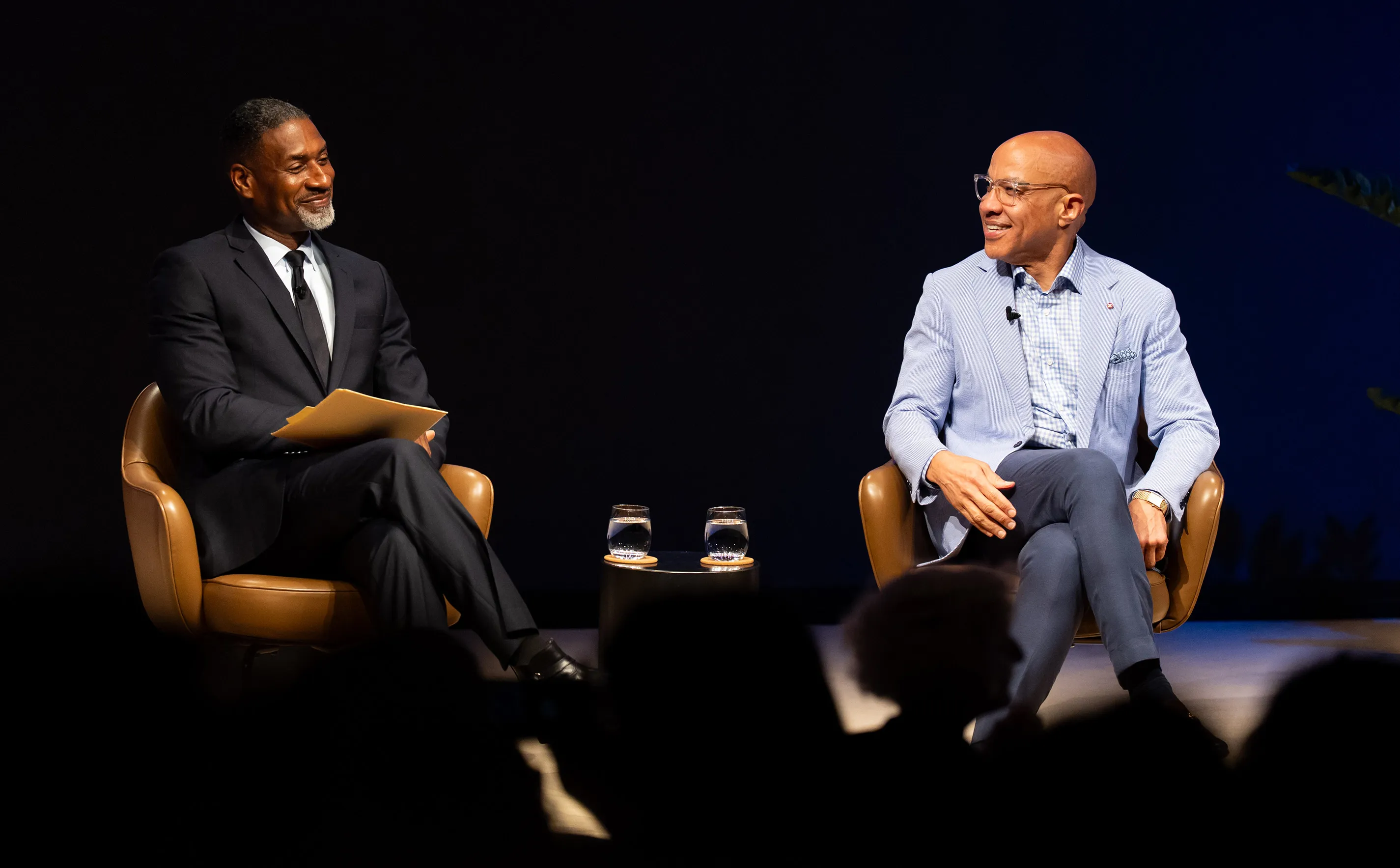 Two men sit in leather chairs on a stage, facing each other and smiling. The man on the left, Charles Blow, is in a dark suit, holding papers. The man on the right, Darren Walker, wears a light blue jacket and glasses.