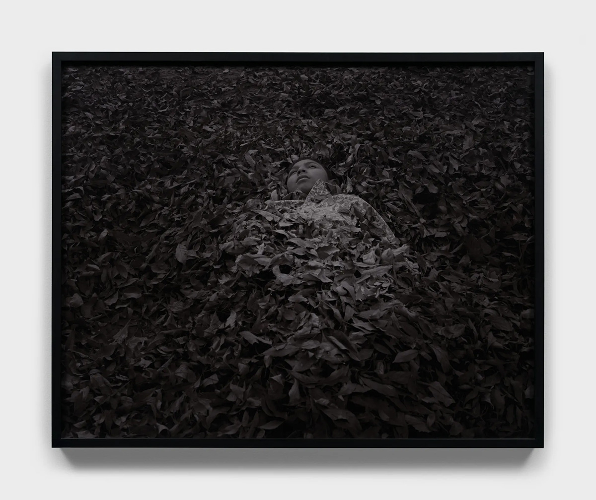A grey-scaled photograph of a person lying in a pile of leaves with only their head and shoulders exposed.