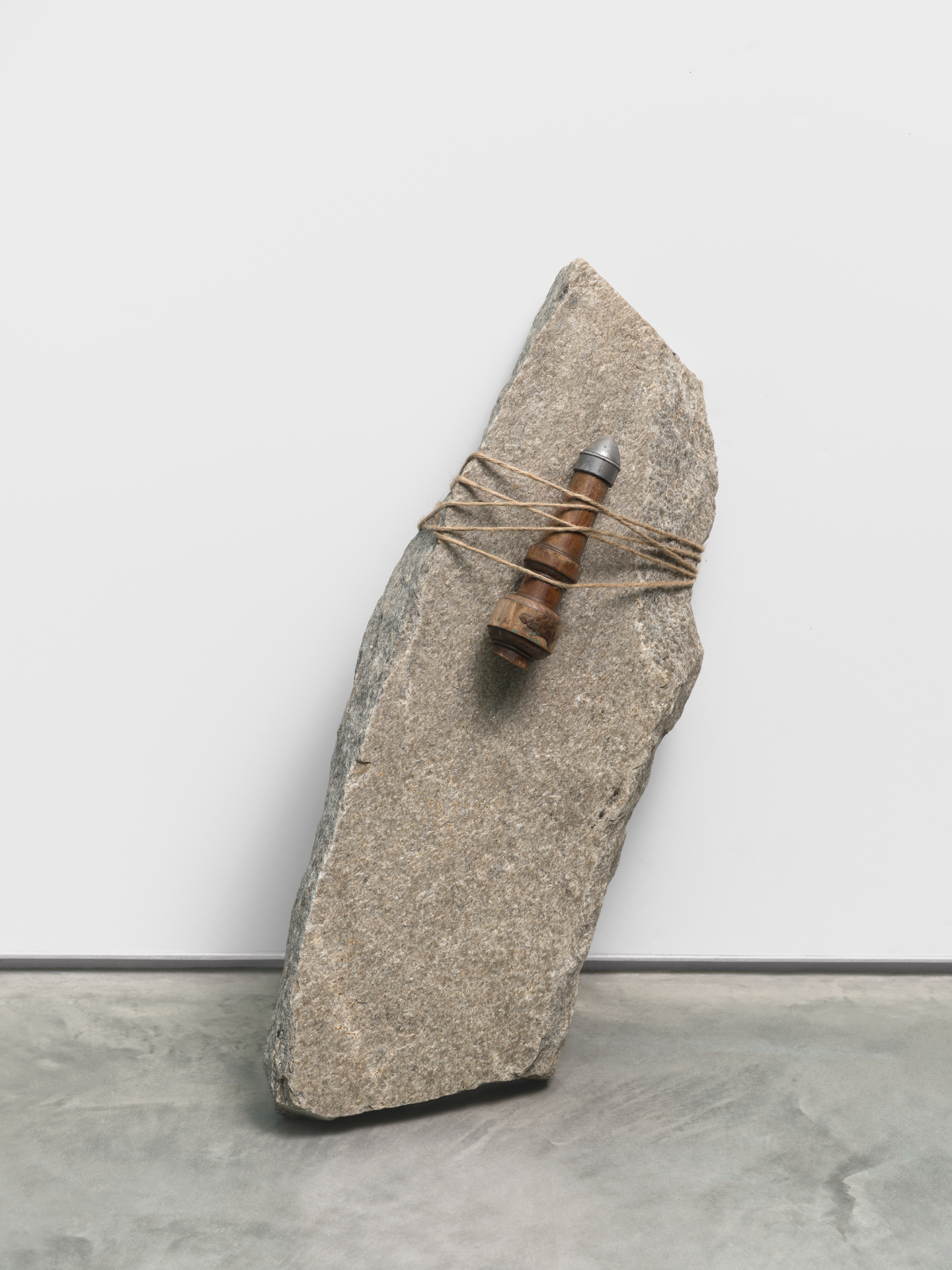 A sculpture of a phallic metal object attached to a slanted, jagged rock by a length of twine which wraps around them both.