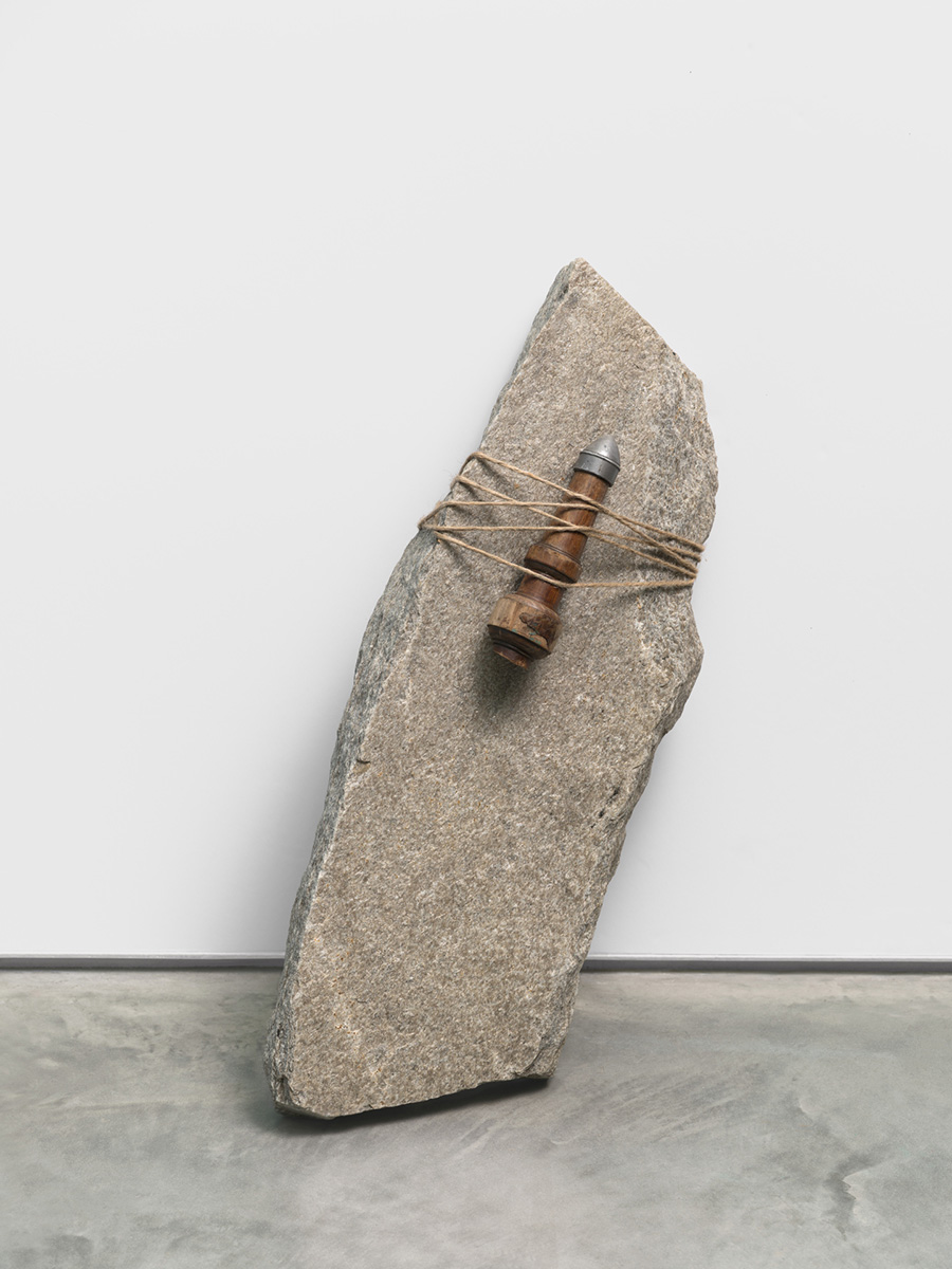 A sculpture of a phallic metal object attached to a slanted, jagged rock by a length of twine which wraps around them both.