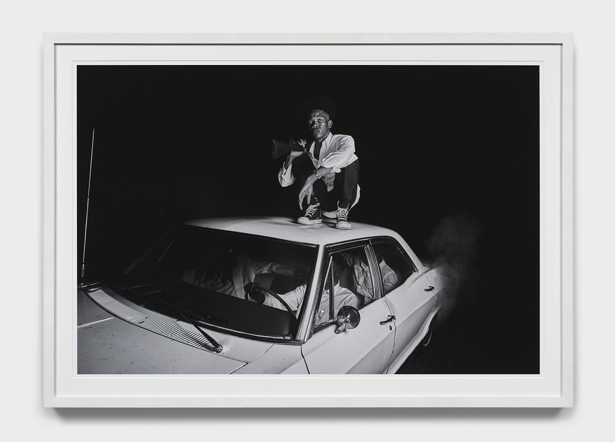 A framed grey-scaled photograph of a Black man squatting on top of a car, resting a rifle on his shoulder. Three people are seated in the car below him. It is a dark night, and the light only illuminates the subjects.