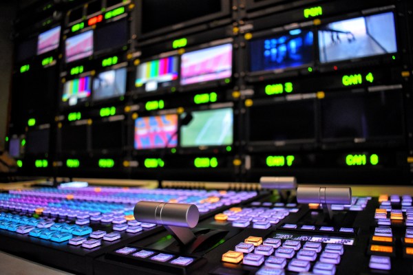 A low-angle shot of a broadcast control room. The foreground shows a large control board with many illuminated buttons and levers, and the background is a wall of monitors displaying various video feeds.