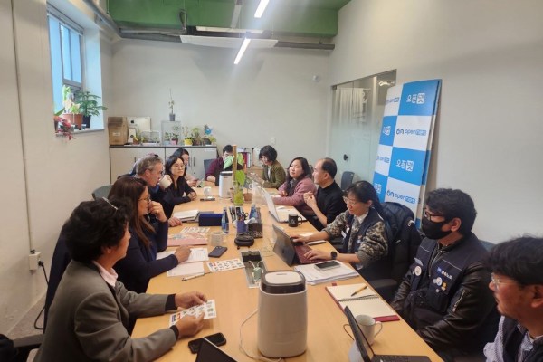 A diverse group of people sits around a large conference table in an office, engaged in a meeting. Laptops, notebooks, and coffee mugs are on the table. A banner with Korean writing is in the background.