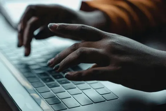 A close-up shot of a person's hands typing on a laptop keyboard. The lighting is low, highlighting the hands and the keys, with a warm-toned sleeve visible in the background.