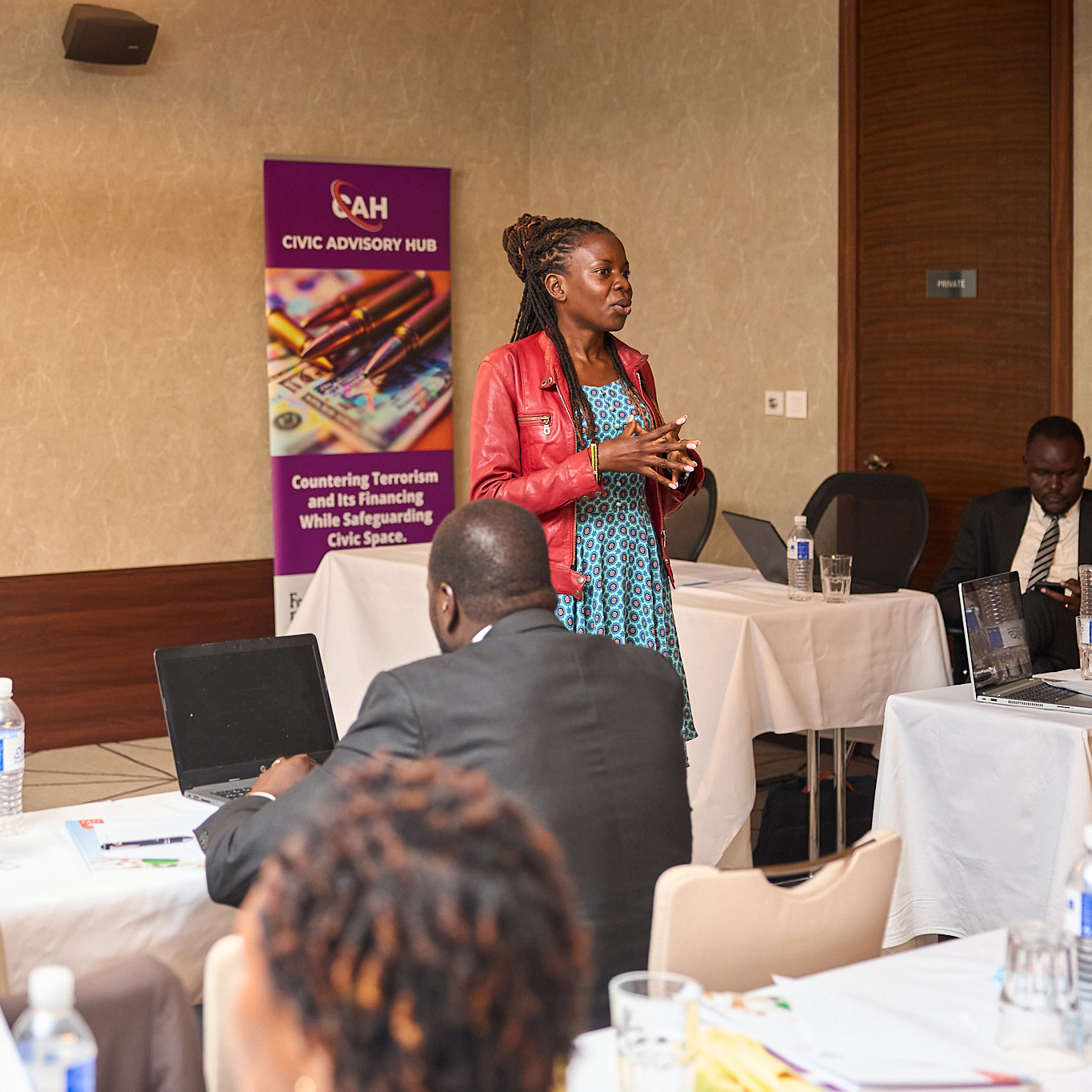 A woman in a red leather jacket and blue patterned dress is speaking at a podium. A projector screen and a banner with the logo "CAH CIVIC ADVISORY HUB" are behind her, and an audience is seated at tables with laptops.