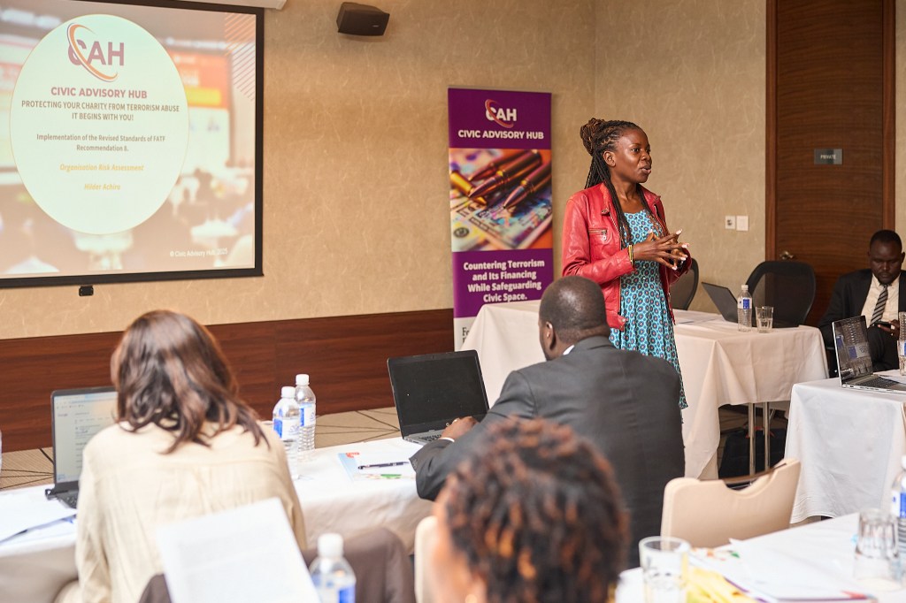 A woman in a red leather jacket and blue patterned dress is speaking at a podium. A projector screen and a banner with the logo "CAH CIVIC ADVISORY HUB" are behind her, and an audience is seated at tables with laptops.