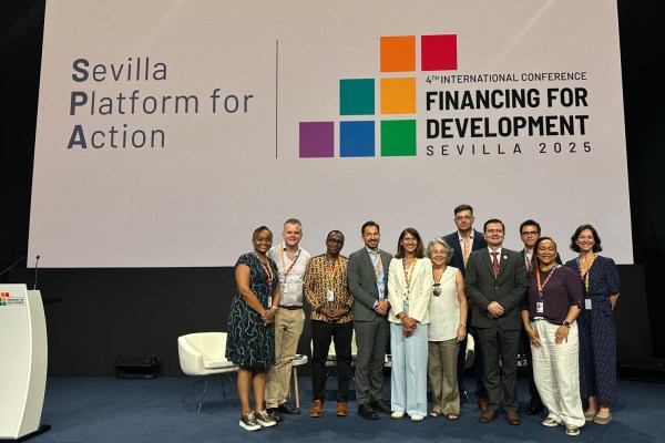 A diverse group of men and women stand smiling on a stage, posing for a photo. A large screen behind them reads "Sevilla Platform for Action" and "4th International Conference on Financing for Development, Sevilla 2025."