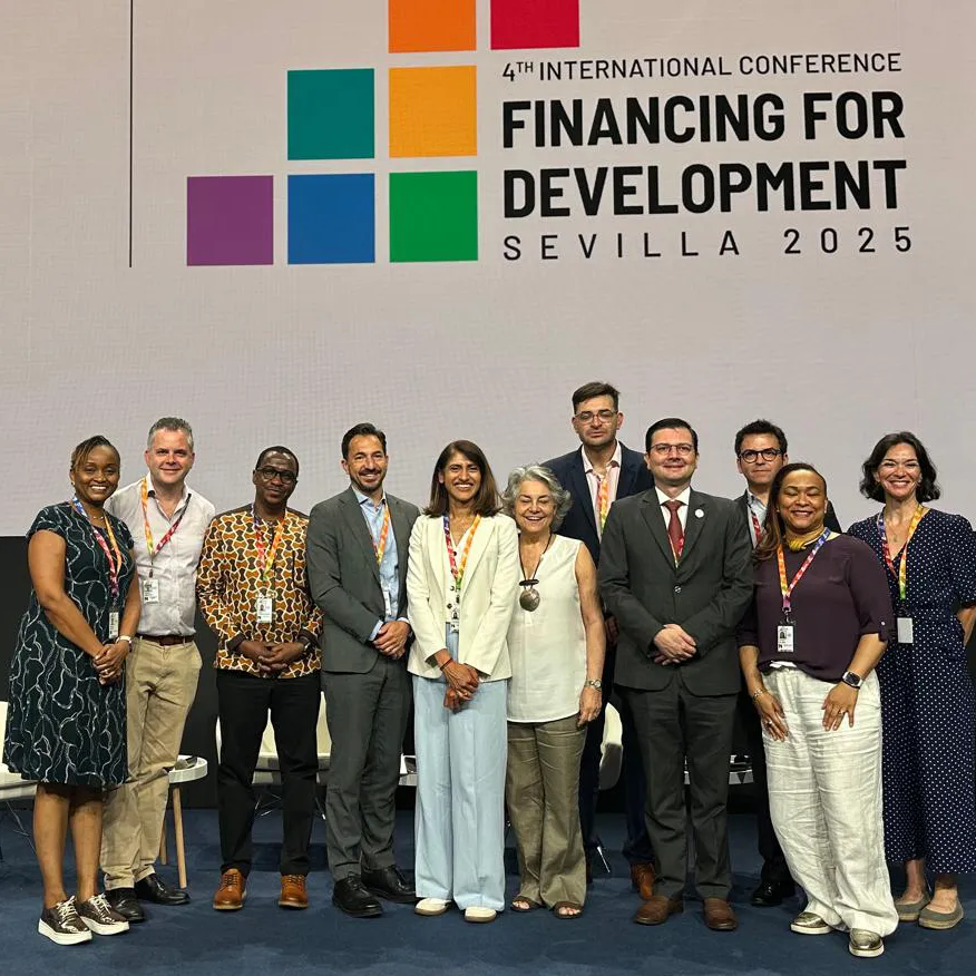 A diverse group of men and women stand smiling on a stage, posing for a photo. A large screen behind them reads "Sevilla Platform for Action" and "4th International Conference on Financing for Development, Sevilla 2025."