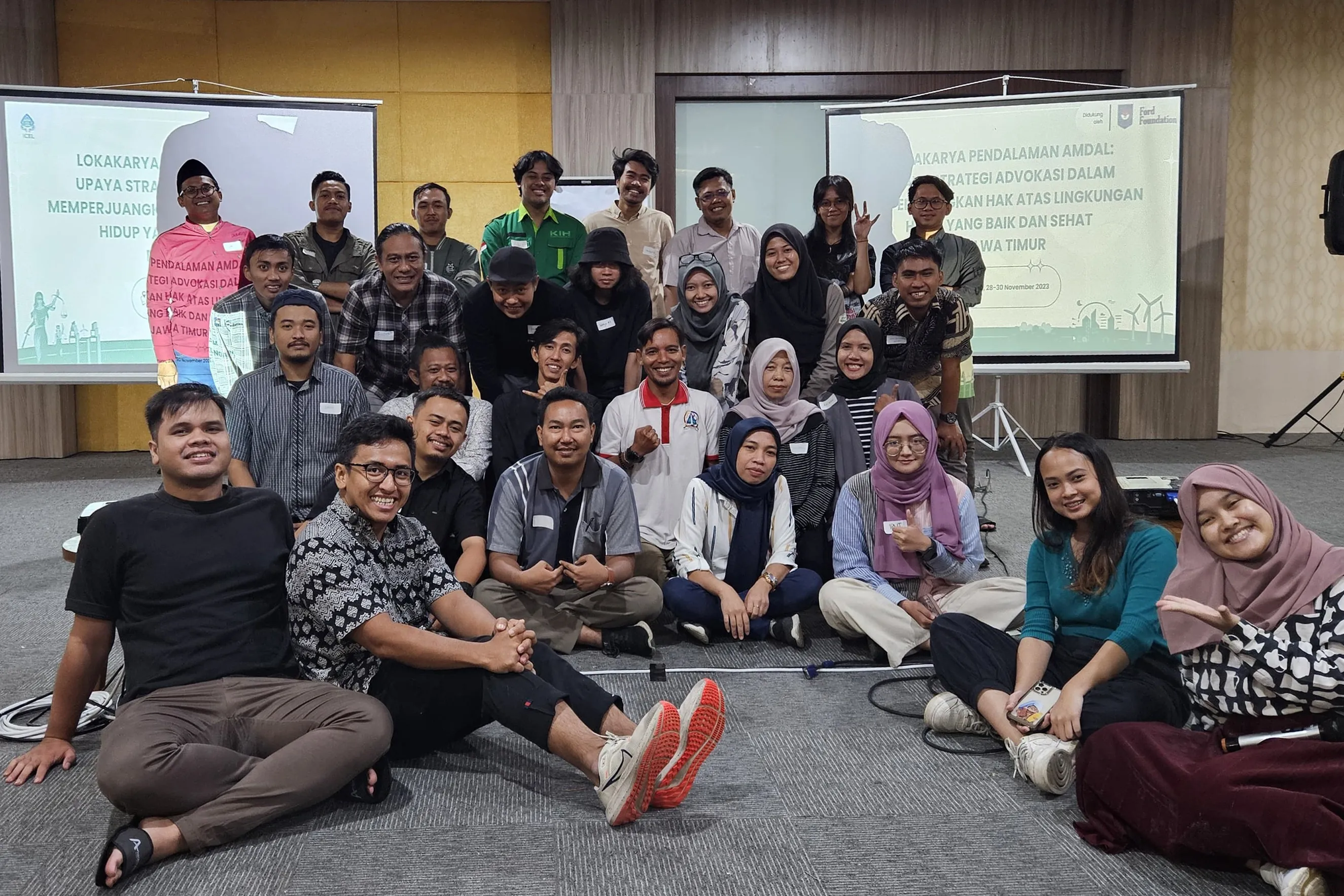 A large group of smiling people sits and stands for a photo in front of two projection screens. The screens show text in a foreign language. The group appears to be at a workshop or conference.