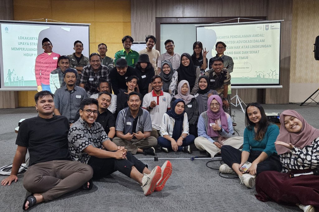 A large group of smiling people sits and stands for a photo in front of two projection screens. The screens show text in a foreign language. The group appears to be at a workshop or conference.