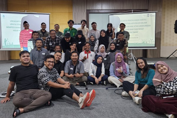 A large group of smiling people sits and stands for a photo in front of two projection screens. The screens show text in a foreign language. The group appears to be at a workshop or conference.