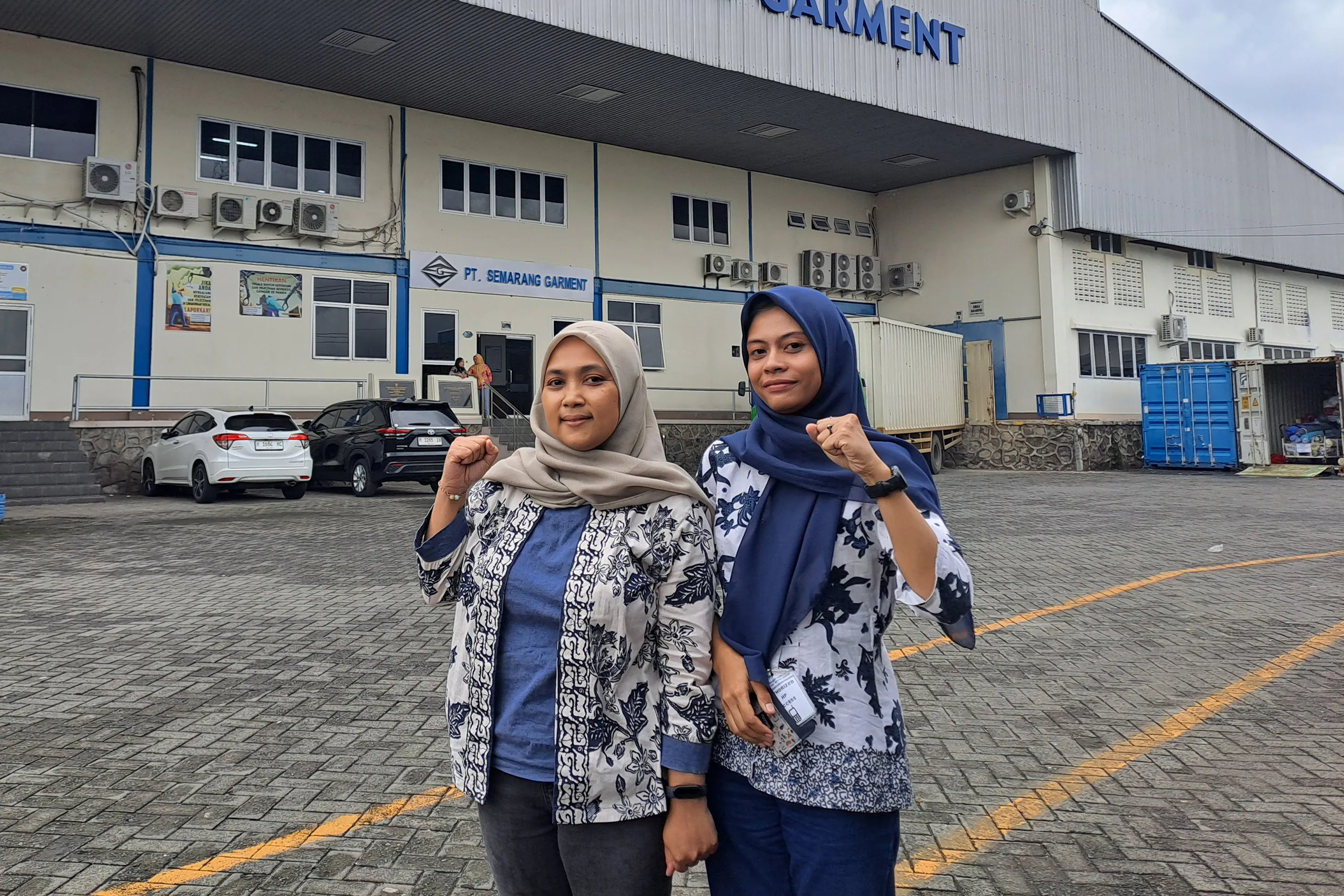 Two women wearing headscarves and patterned jackets stand outdoors in front of a building with a sign that reads "PT. SEMARANG GARMENT." They are smiling and raising a clenched fist in a show of solidarity.