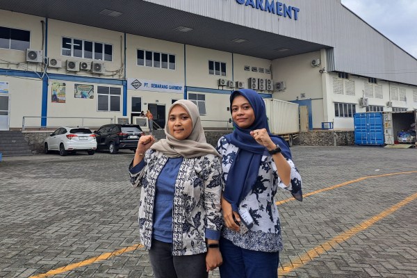 Two women wearing headscarves and patterned jackets stand outdoors in front of a building with a sign that reads "PT. SEMARANG GARMENT." They are smiling and raising a clenched fist in a show of solidarity.