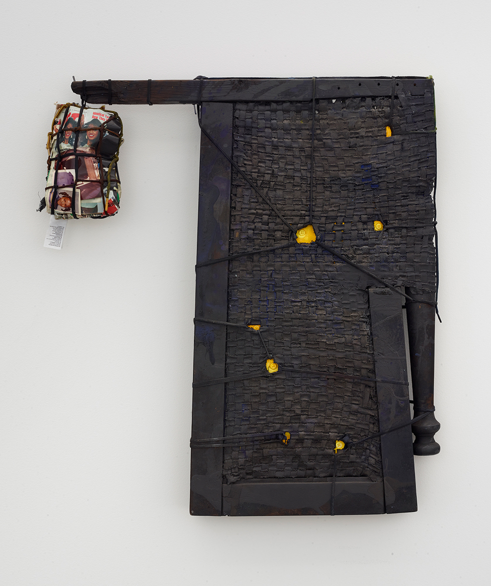 A mixed-media sculpture of discarded furniture amalgamation. There’s an extended arm in which a bundle of magazine clippings of Black women dangle off by thread.