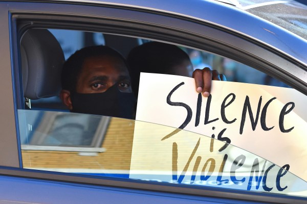 A person wearing a black face mask looks out from the passenger window of a car, holding up a sign that reads "Silence is Violence" in black handwriting against a white background.