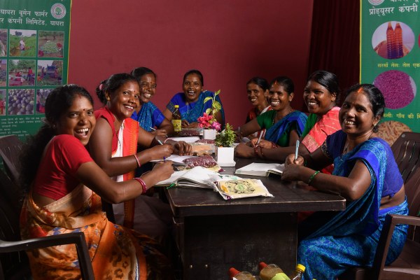 A group of eight women are seated around a rectangular table, smiling and looking towards the camera. Most of the women are holding pens and have open notebooks or papers in front of them. On the table are also small bags of what appear to be dried spices or herbs, and a couple of bottles of oil. The women are dressed in colorful traditional Indian attire. In the background, there are posters on the walls with images of crops and agricultural scenes, along with text in Hindi that mentions "Ghaghra Women Farmer Producer Company Limited."