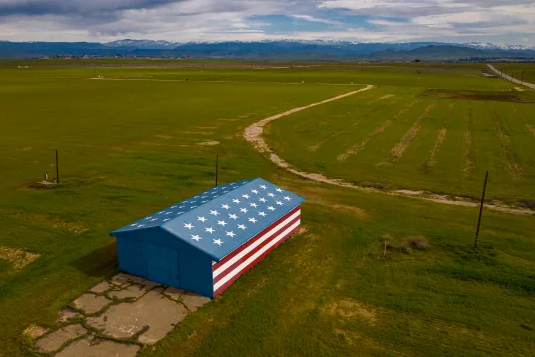 An aerial view of a barn or shed painted with an American flag pattern, situated in the middle of a large green field. The sky is partly cloudy, and a road and mountains are in the distance.