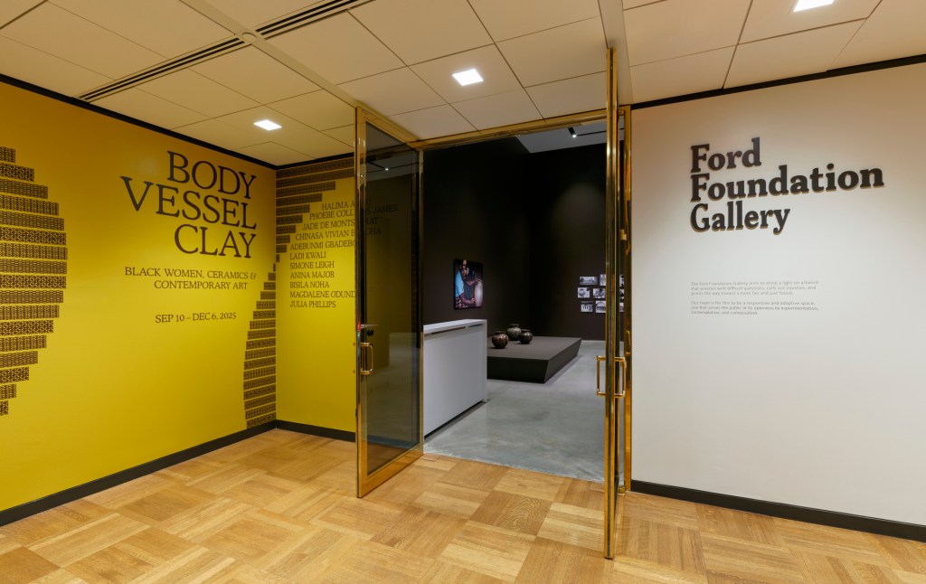 An entrance with doors propped open into a gallery exhibition. The left wall has geometric designs and text that reads “Body Vessel Clay”