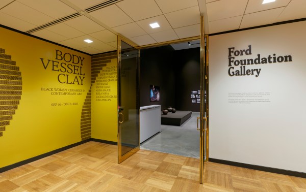 An entrance with doors propped open into a gallery exhibition. The left wall has geometric designs and text that reads “Body Vessel Clay”