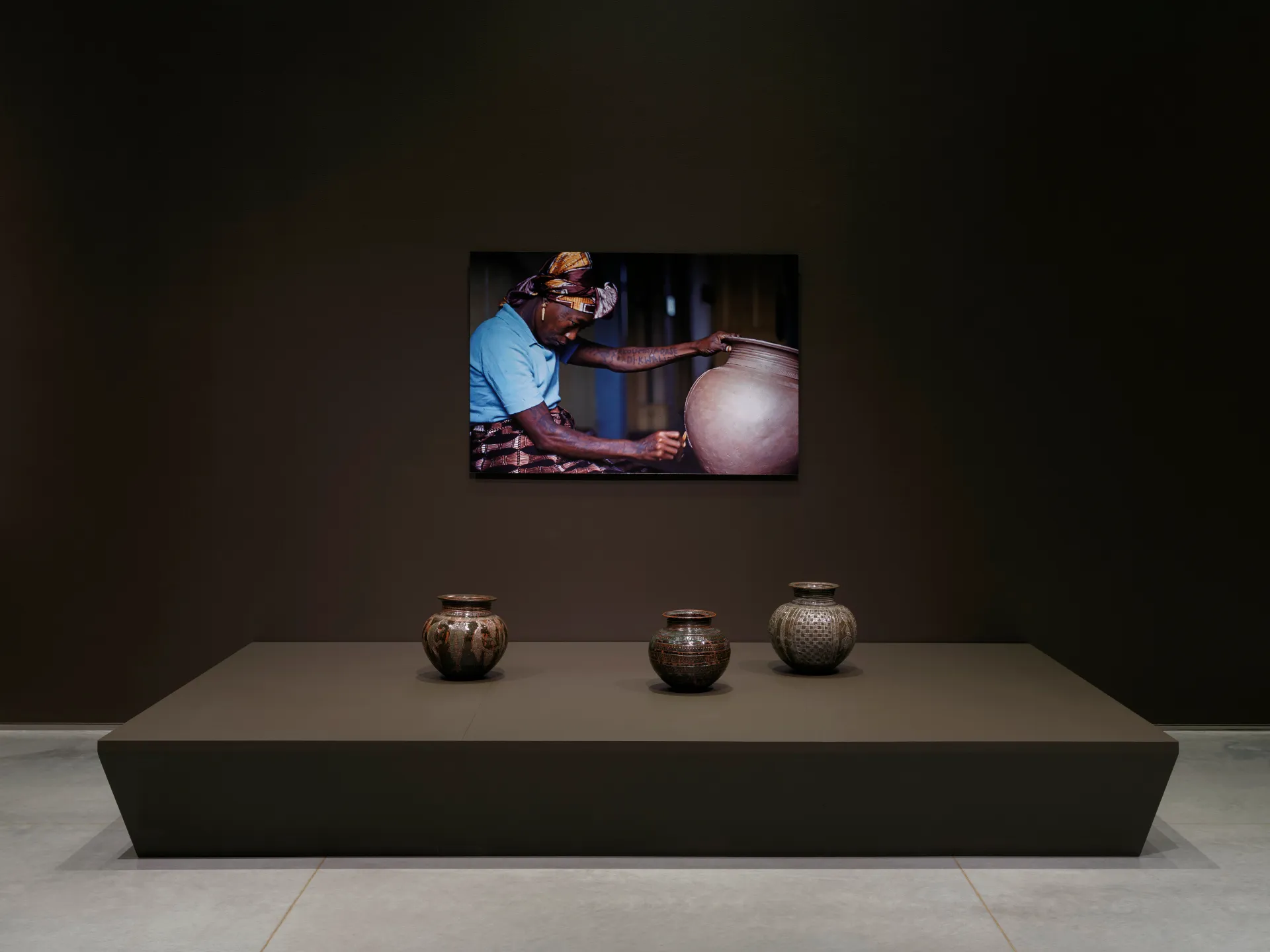 Three glazed pots below a mounted photograph of a Black woman carving into a clay pot.