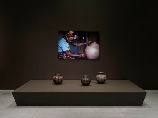 Three glazed pots below a mounted photograph of a Black woman carving into a clay pot.