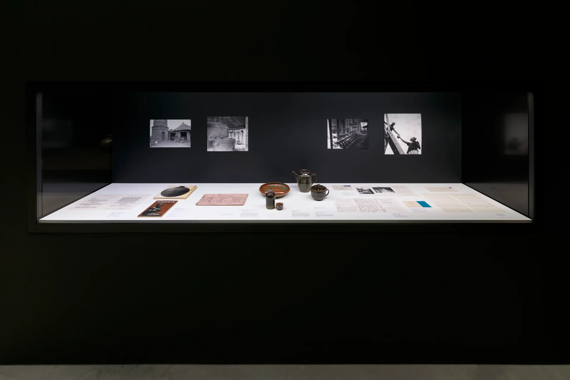 A glass vitrine holding archival paper objects, ceramic works, and historic photography documentation of a pottery center.
