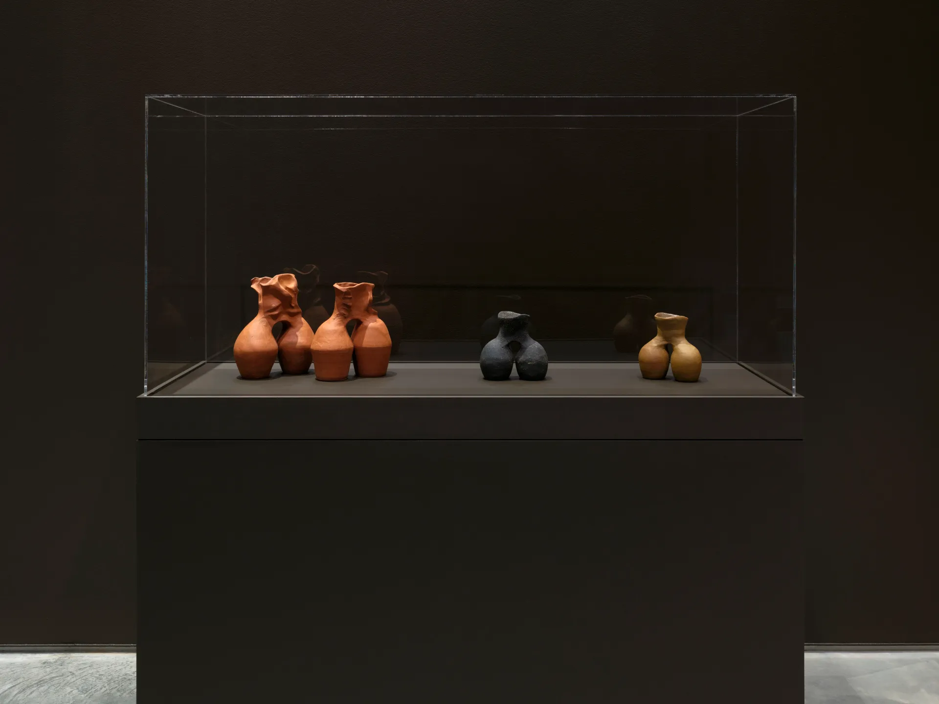 A glass vitrine holding four ceramic sculptures that each have a similar two-barrel shape, but made of different ceramic material and of slightly different heights.
