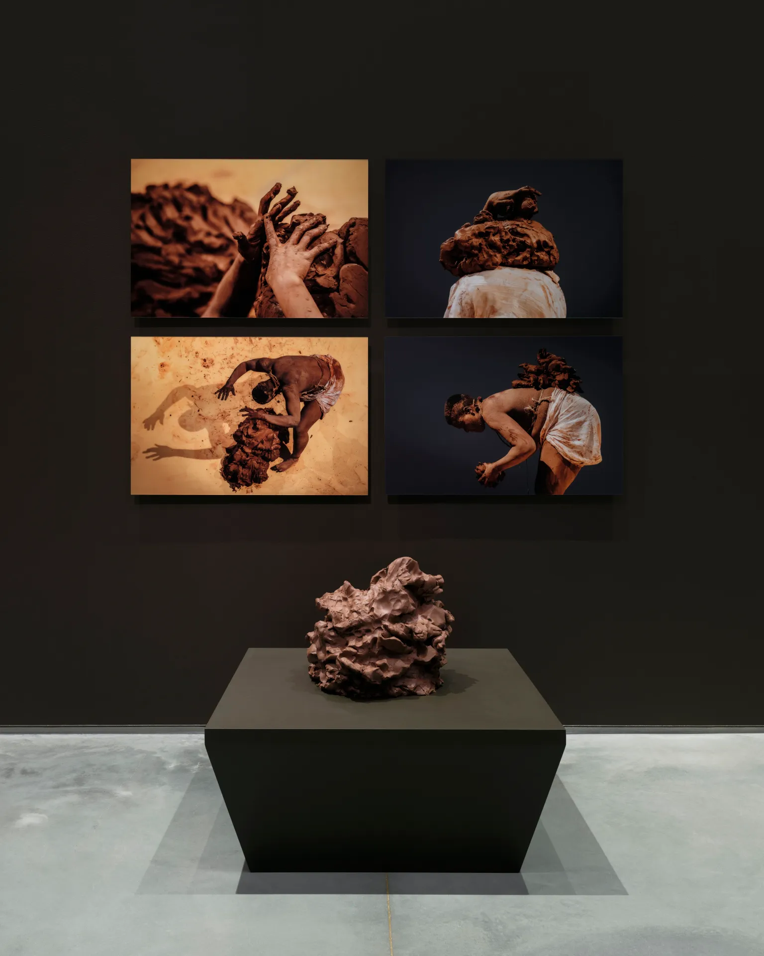 An installation composed of four wall-mounted photos in a grid with a pedestal piled with a large mound of rust-colored clay.