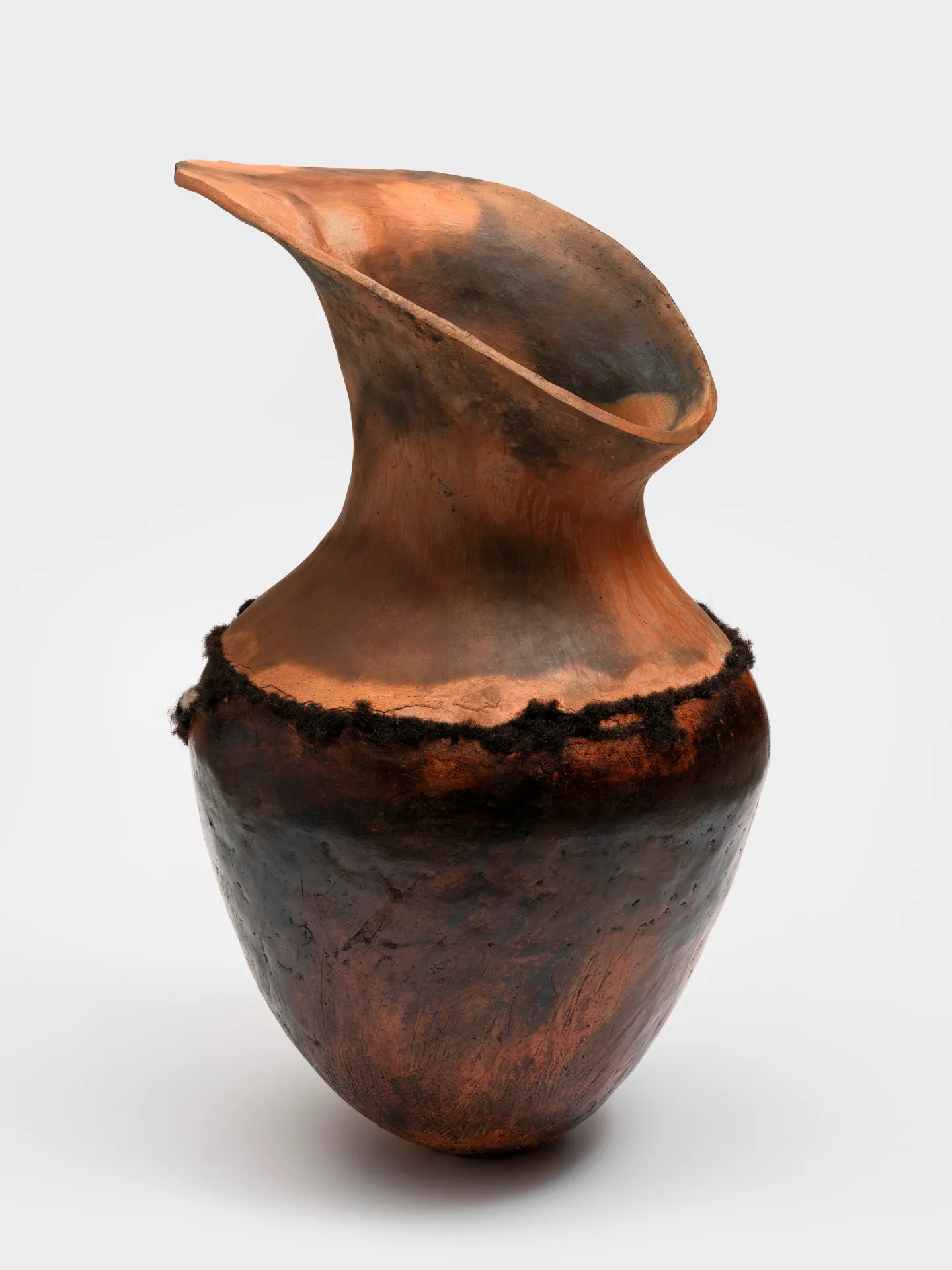 A large pit-fired ceramic pot with a wide teardrop-shaped mouth and dark curly hair belted around the vessel.