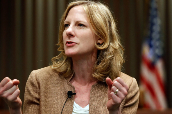 A candid photo of Heather Gerken with shoulder-length blonde hair, wearing a tan blazer over a white top. She is speaking passionately, holding both fists slightly raised. A blurred American flag is visible in the background.