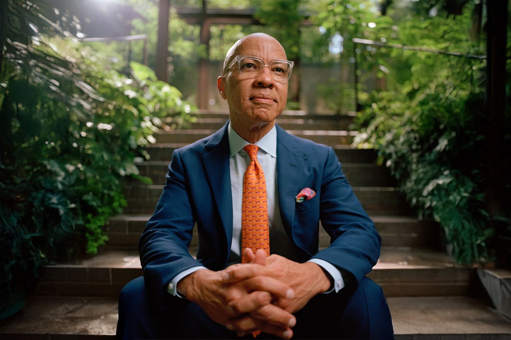 Darren Walker sitting on steps of the Ford Foundation atrium steps. He’s wearing a blue suit, light blue shirt and orange tie.
