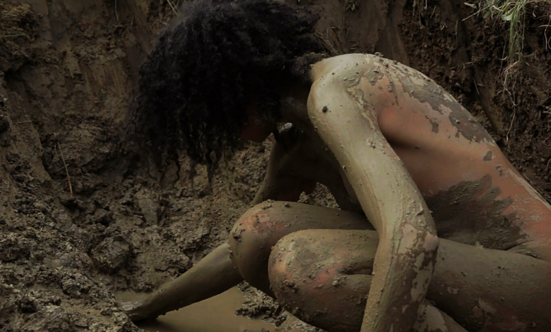 A screenshot of a 9-minute video of a naked light-skinned Black woman in a dirt hole in an open field digging and bathing in mud.