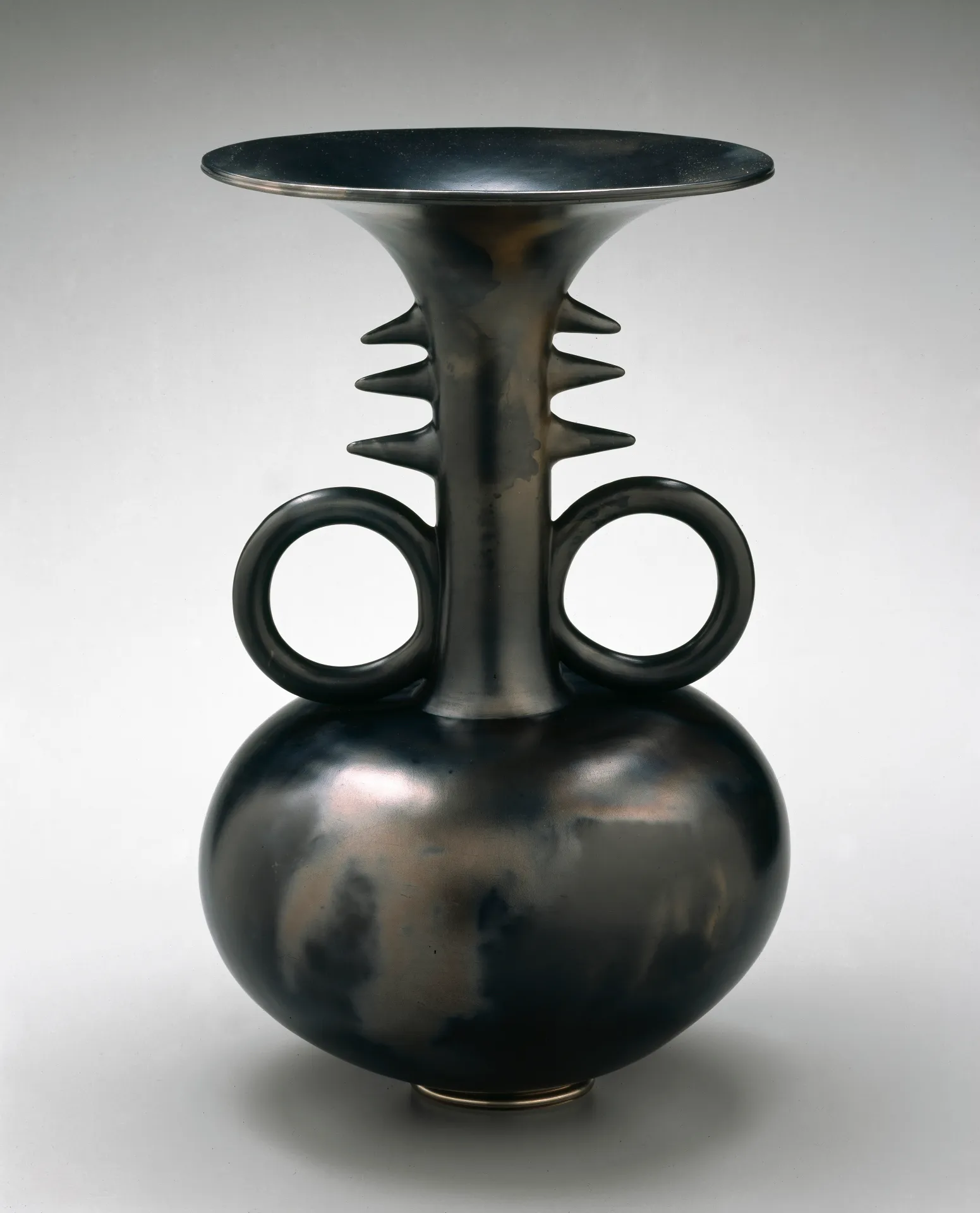 A black ceramic vase with a body in the shape of a horizontal oval narrowing into a neck with circular handles and spikes on either side and opening into a wide, flat mouth.