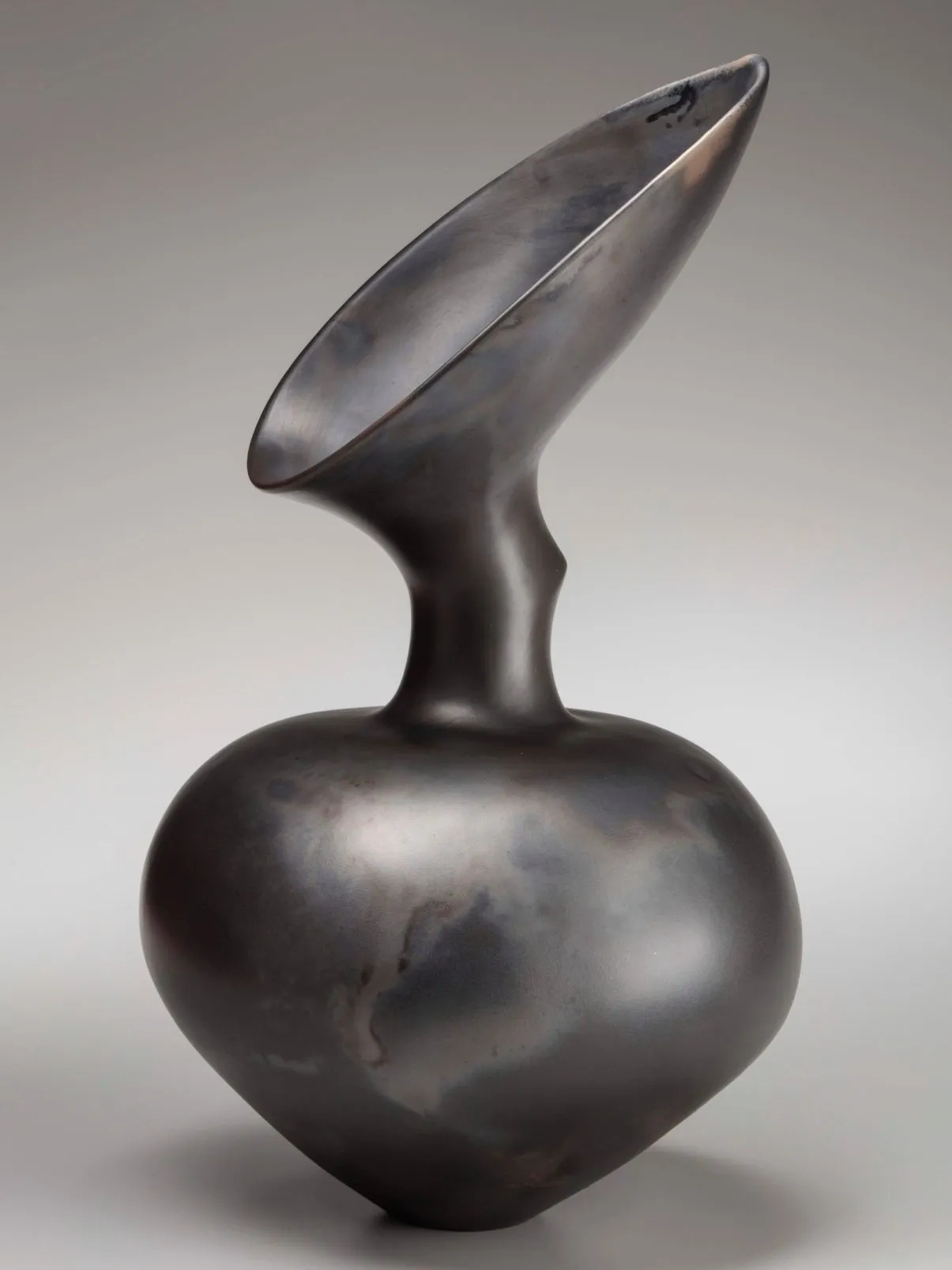 A black ceramic vase with a heart-shaped body meeting at a thin, pointy neck and opening into a large teardrop-shaped mouth slanted upwards.