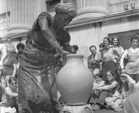 A Black woman shaping a vertical, clay vase in front of an audience.