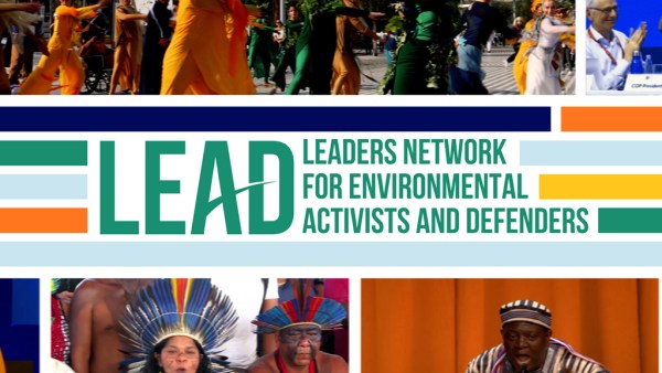 A vibrant crowd of people in traditional dress (yellow, green, and orange) and speakers on stage, surrounding the text "LEADERS NETWORK FOR ENVIRONMENTAL ACTIVISTS AND DEFENDERS".