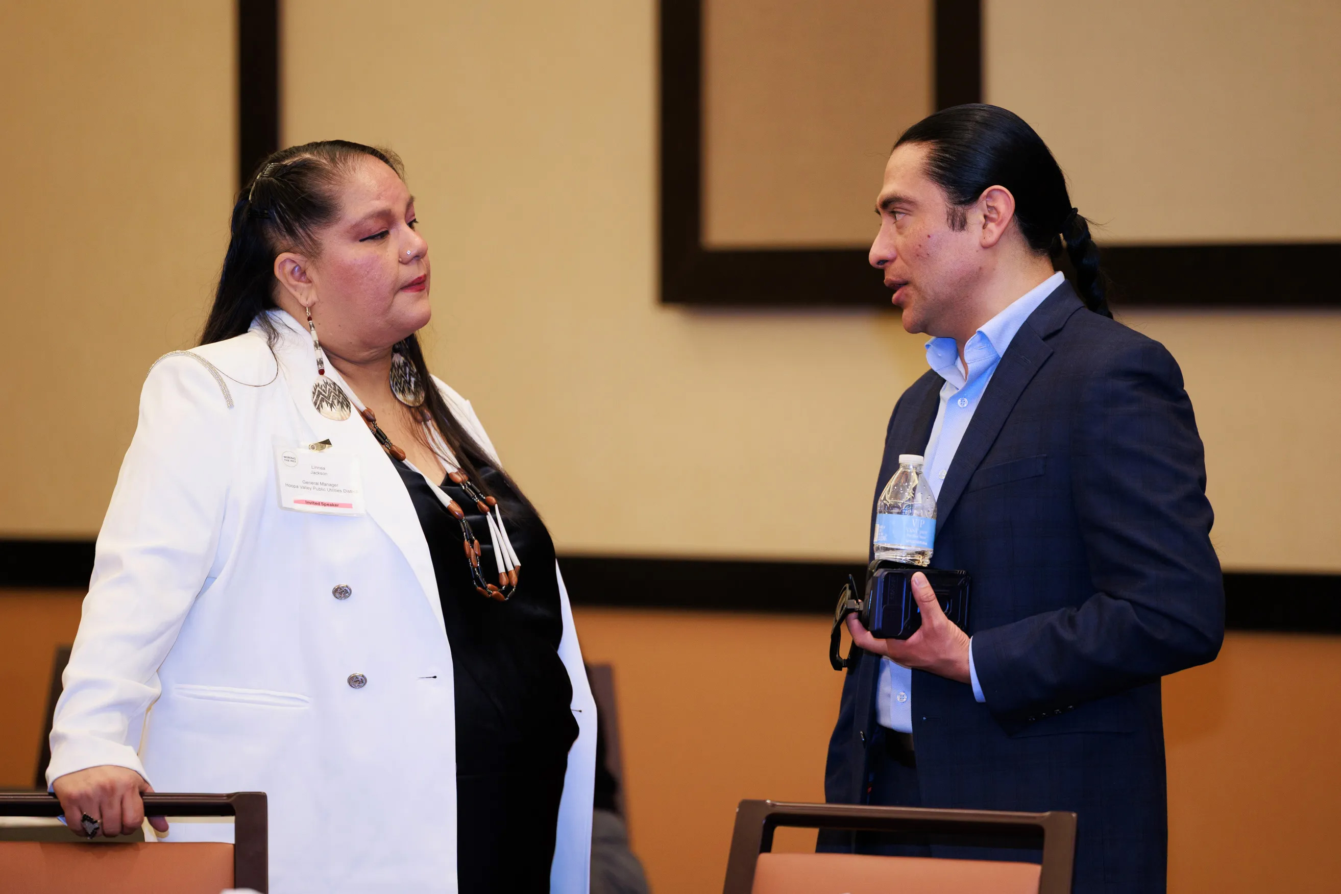 Two Indigenous leaders converse during a conference. The woman wears a white blazer and beaded jewelry; the man wears a blue suit and holds a water bottle, discussing Tribal Digital Sovereignty.