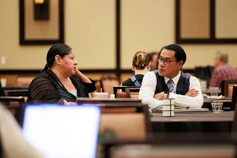 Two conference attendees (an Indigenous woman and a man in a vest and tie) in serious discussion at a table. Image context: Tribal Digital Sovereignty, broadband access, digital equity.