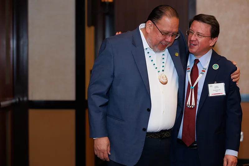 Two men, one Indigenous leader wearing a turquoise necklace and the other a suit and tie, embrace at a conference. Focus keywords: Tribal sovereignty, digital inclusion, partnerships, community development.