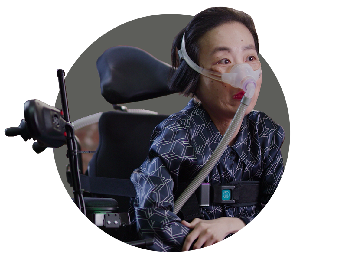 Alice Wong, an Asian American woman in a wheelchair wearing a mask over her nose attached to a tube for a BiPAP machine that helps her breathe. She is wearing a navy striped shirt and dark pants.