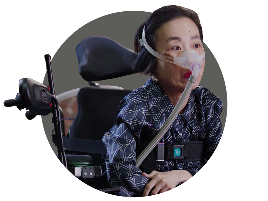 Alice Wong, an Asian American woman in a wheelchair wearing a mask over her nose attached to a tube for a BiPAP machine that helps her breathe. She is wearing a navy striped shirt and dark pants.