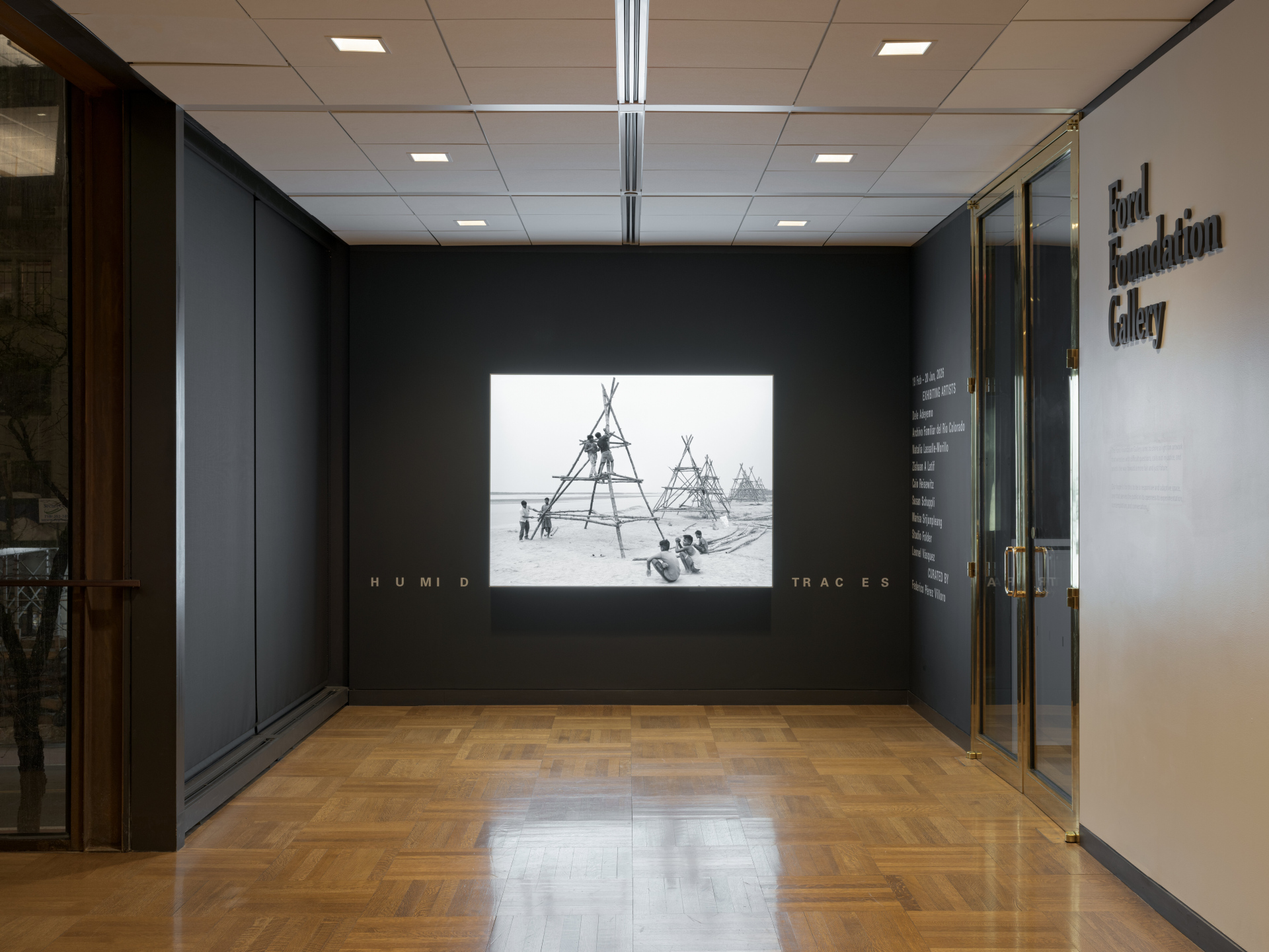 Outside of a gallery entrance, a bright lightbox displays boys assembling a tall bamboo structure on the shore of a beach.