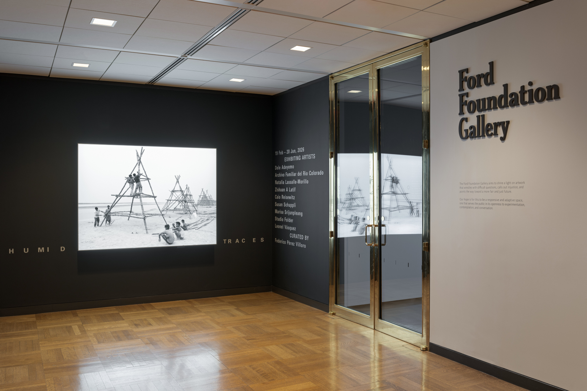 An entrance to the Ford Foundation Gallery exhibition displaying the exhibition title “HUMID TRACES” and the list of artists in a standardized font. A bright lightbox of boys assembling a bamboo structure is hung on the middle wall next to glass doors.