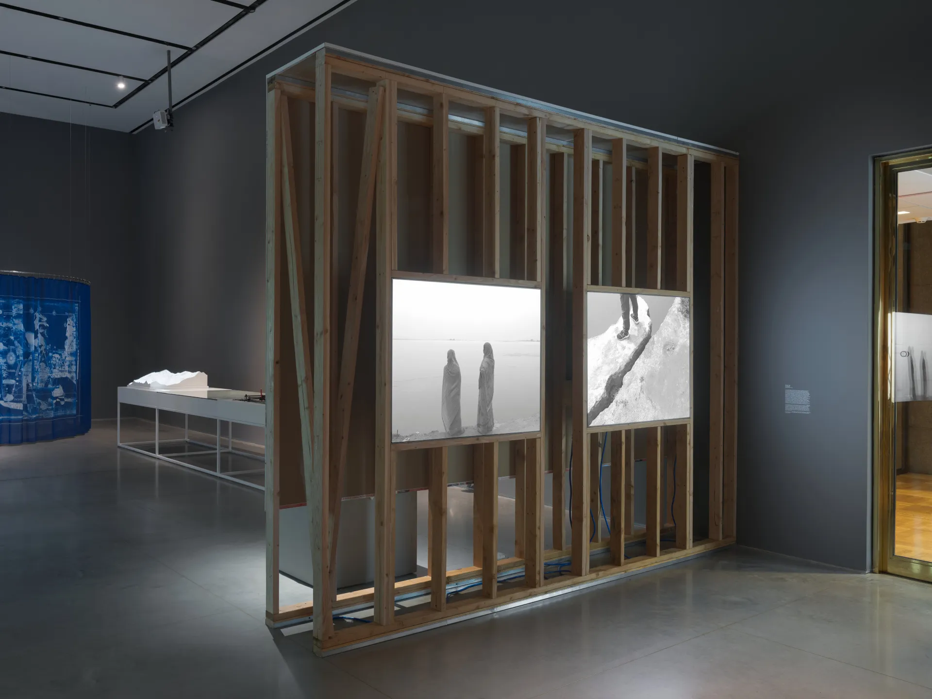 An unfinished wall construction holding two light boxes. One lightbox displays the backs of two women in saris overlooking water. The other shows the legs of someone standing on a cliff’s edge.