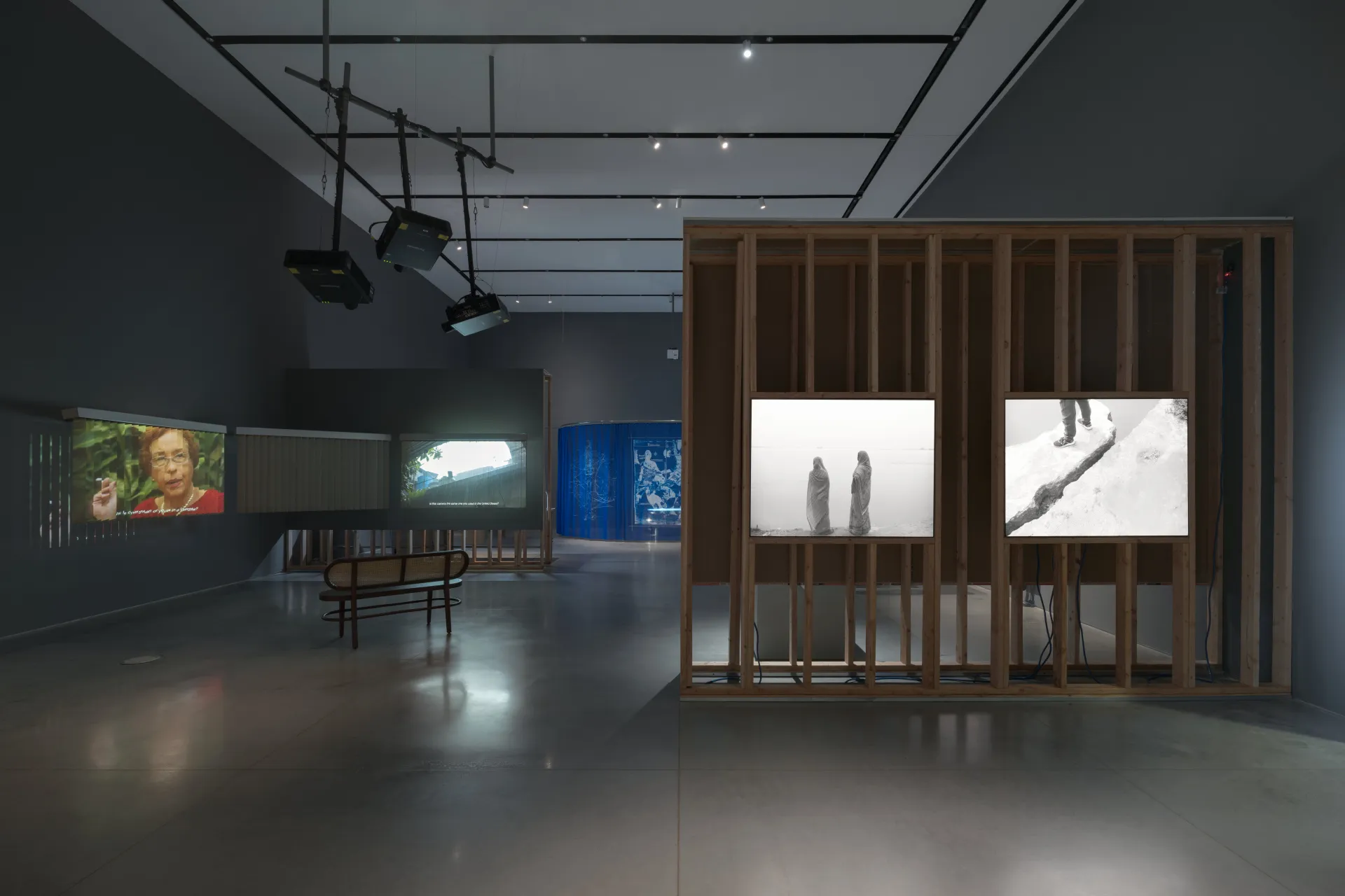 A gallery exhibition showcasing several projections, light boxes within a wall construction, and a blue tapestry in the distance.