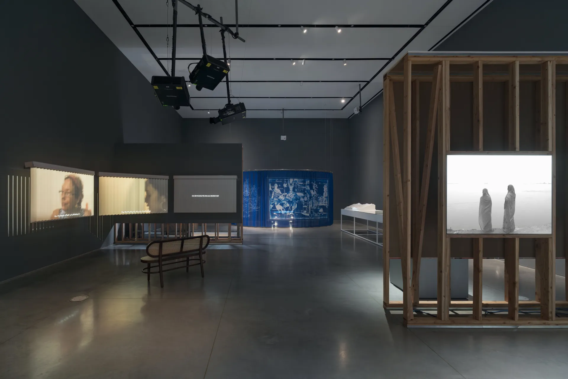 An art exhibition showcasing several projections, a wall construction with a lightbox, and in the distance, a blue tapestry and corner of a metal table.