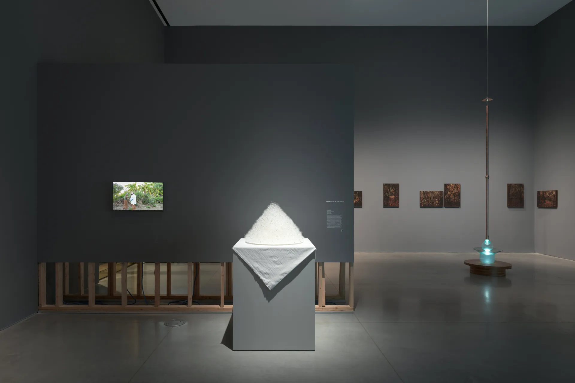 A gallery interior showing a wall with a small video monitor and a white pyramid soft-sculpture atop a pedestal. Behind this wall stands a glass illuminating sculpture against a wall with hanging dark prints.